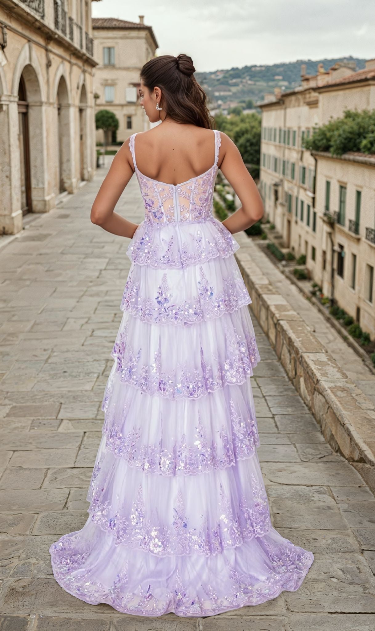 Wearing the Tiered Long Ruffle Prom Dress: Chicas C2433, a woman stands on a stone walkway amid historic buildings and lush greenery.